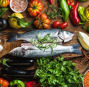 Raw uncooked seabass fish with vegetables, grains, herbs and spices on chopping board over rustic wooden background